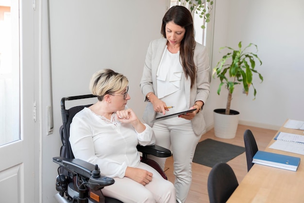 Una mujer de negocios con discapacidad sentada en silla de ruedas reunida con su compañero de trabajo usando una tableta digital