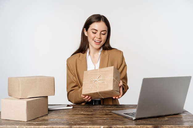 Mujer de negocios, dueña de una tienda sentada cerca de una laptop, sosteniendo una caja con el pedido del cliente y sonriendo, trabajando en una computadora, fondo blanco