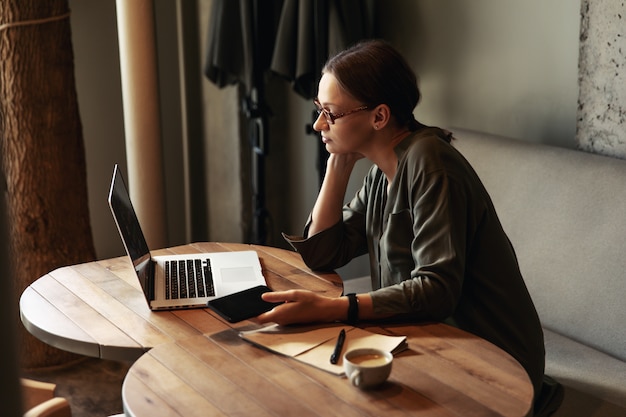 Mujer de negocios enfocada en gafas elegantes sentarse en la cafetería trabajando en la computadora portátil, concentrada mujer seria trabajando con la computadora