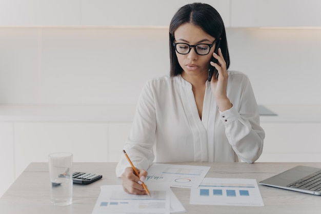 Una mujer de negocios enfocada habla por teléfono para discutir el análisis financiero del proyecto y escribir una nota en el escritorio de la oficina