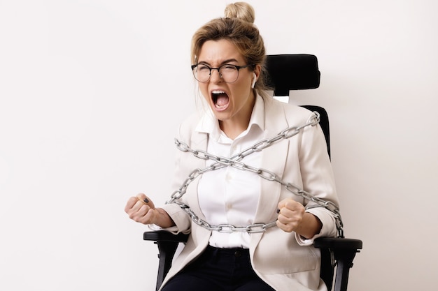 Mujer de negocios enojada atada a la silla de oficina con cadenas. Concepto de deudas o horas extraordinarias.