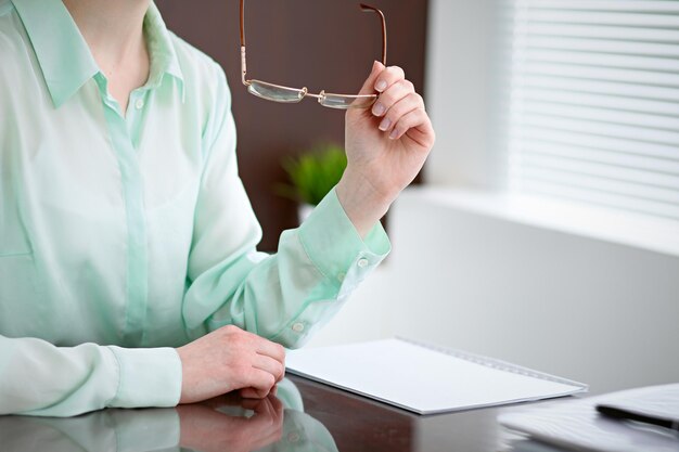 Mujer de negocios entrega una blusa verde sentada en el escritorio de la oficina y sosteniendo gafas, la ventana derecha. Ella está pensando en los problemas de negocios, los éxitos, los conceptos