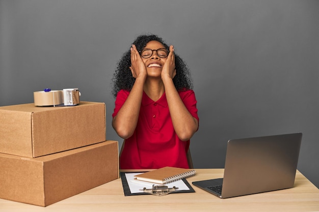 Mujer de negocios de entregas con inventario y cajas se ríe alegremente manteniendo las manos en la cabeza