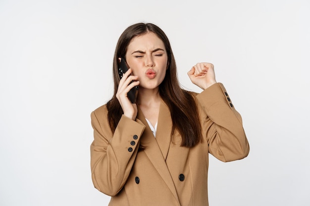 Mujer de negocios entusiasta hablando por teléfono móvil, reaccionando sorprendida y feliz de llamar, recibir buenas noticias, de pie sobre fondo blanco