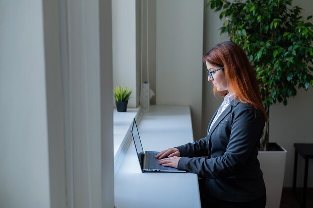 Mujer de negocios escribiendo en la computadora portátil mientras está sentada en un café junto a la ventana Una gerente con traje trabaja de forma remota durante un descanso detrás de la barra