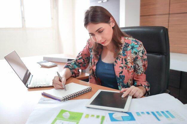 Mujer de negocios escribiendo en un libro mientras usa una tableta digital en la mesa