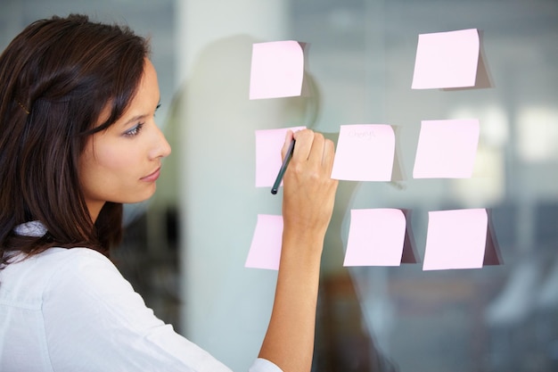 Mujer de negocios escribiendo y planificando una estrategia de lluvia de ideas o ideas en un tablero de vidrio en la oficina Empleada que trabaja en tareas para una nota adhesiva recordatoria o un plan de proyecto en el lugar de trabajo