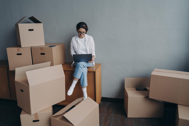 Mujer de negocios española alquila apartamento Niña feliz está sentada entre cajas y trabajando en una computadora portátil