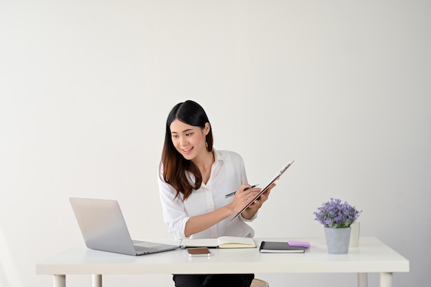 Una mujer de negocios está tomando notas en papel en un portapapeles mientras mira la pantalla de su computadora portátil