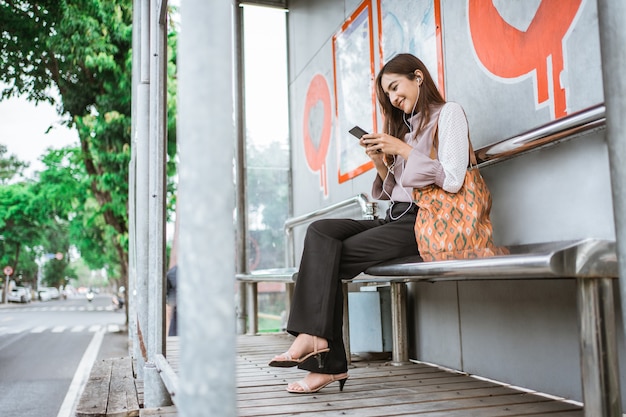 Mujer de negocios en una estación de autobuses esperando que llegue el autobús mientras usa el teléfono