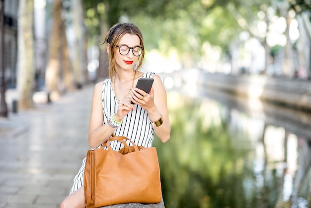 Mujer de negocios con estilo joven sentada con teléfono y bolso al aire libre en el hermoso parque con río durante el día soleado