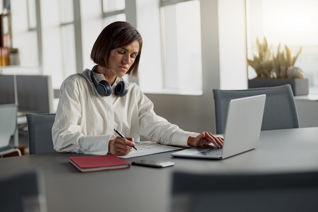 Mujer de negocios europea enfocada que trabaja con una computadora portátil y toma notas mientras está sentada en una oficina moderna