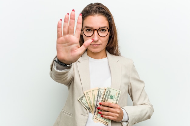 Mujer de negocios europea joven que sostiene los billetes de dólar que se colocan con la mano extendida que muestra la señal de stop, previniéndole.