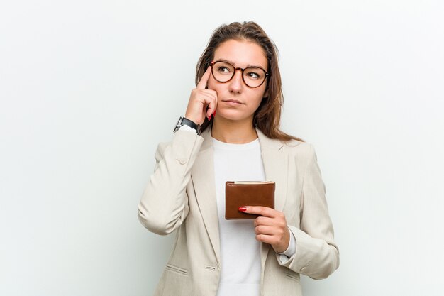 Mujer de negocios europea joven que sostiene una cartera que señala su sien con el dedo, pensando, centrado en una tarea.