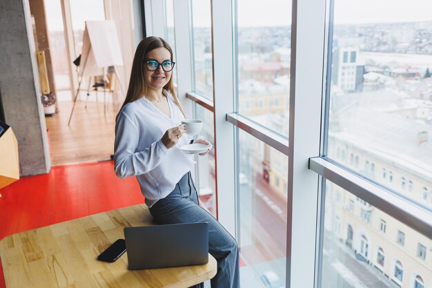 Mujer de negocios europea que usa una computadora portátil mientras trabaja El concepto de una mujer exitosa moderna Joven hermosa mujer sonriente con café sentado en un café Interior de la oficina abierta