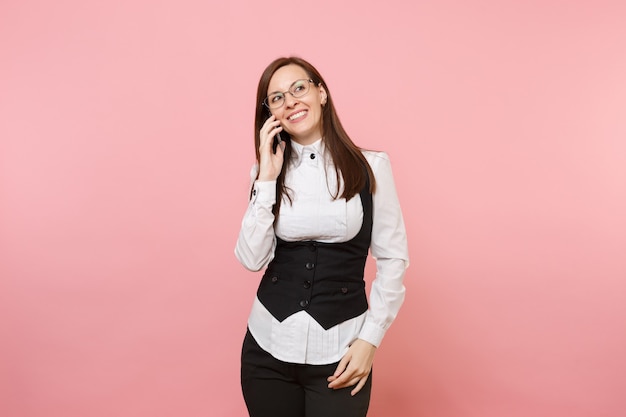 Mujer de negocios exitosa joven en traje negro y gafas sosteniendo y hablando por teléfono móvil aislado sobre fondo rosa pastel. Jefa. Riqueza de carrera de logro. Copie el espacio para publicidad.