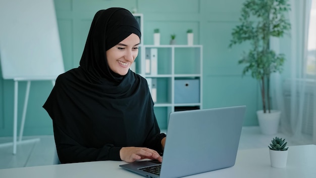 Mujer de negocios exitosa musulmana en hijab mira la computadora portátil sonriendo trabajando en línea estudiando