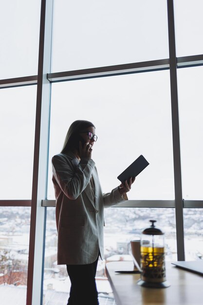 Mujer de negocios experimentada sosteniendo un teléfono mientras está de pie en el interior de una oficina moderna cerca de una gran ventana con vista al paisaje urbano La ejecutiva femenina se ve complacida después de una reunión exitosa