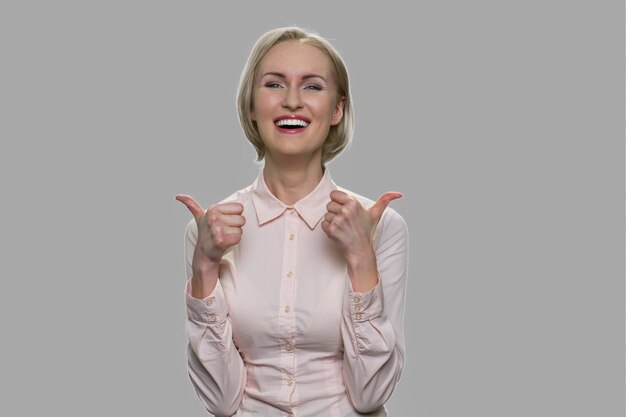 Mujer de negocios feliz dando dos pulgares para arriba. Mujer rubia alegre en camisa formal gesticulando el pulgar hacia arriba con ambas manos sobre fondo gris.