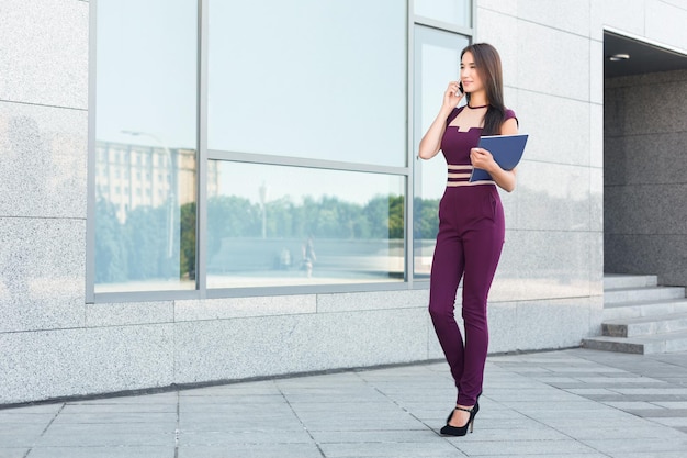 Mujer de negocios feliz hablando por móvil y trabajando con documentos. Mujer trabajando con papeles y consultando en un teléfono inteligente, de pie cerca del moderno centro de oficinas, copiando espacio