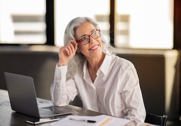 Mujer de negocios feliz posando con anteojos sentado en la computadora portátil en la oficina