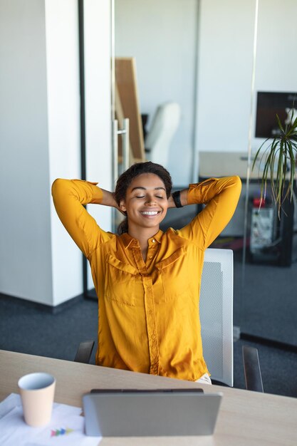 Mujer de negocios feliz y relajada cogida de la mano detrás de la cabeza sentada en el escritorio y descansando trabajando en la oficina moderna recortada