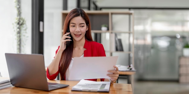 Mujer de negocios feliz sentada en el escritorio detrás de su computadora portátil y hablando con alguien en su teléfono móvil mientras trabaja en la oficina