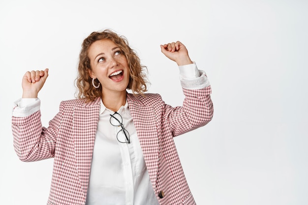 Mujer de negocios feliz en traje saltando y haciendo gesto de bomba de puño, riendo y divirtiéndose, de pie bombeada contra fondo blanco