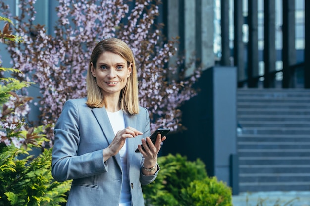 Mujer de negocios feliz usa el teléfono lee el mensaje mirando la pantalla del teléfono inteligente se regocija y sonríe fuera de la oficina