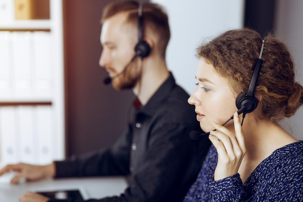 Foto mujer de negocios hablando por auriculares mientras se sienta con un colega de barba roja en una oficina moderna y soleada. grupo de personas diversas en el centro de llamadas.