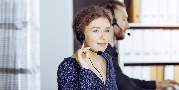 Mujer de negocios hablando por auriculares mientras se sienta con un colega de barba roja en una oficina soleada. Grupo de personas diversas en call center