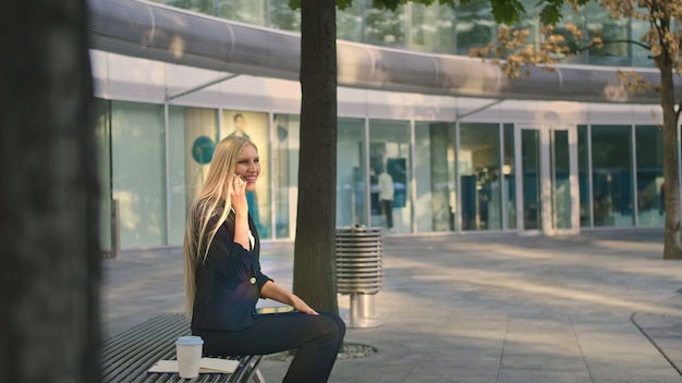 Mujer de negocios hablando por teléfono y saludando con la mano. Elegante jovencita moderna sentado en el banco y saludando con la mano a un amigo mientras habla en el teléfono inteligente al aire libre.