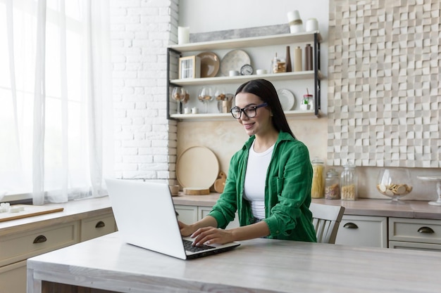 Mujer de negocios hermosa joven en gafas y camisa verde trabajando en casa en la cocina en la computadora portátil