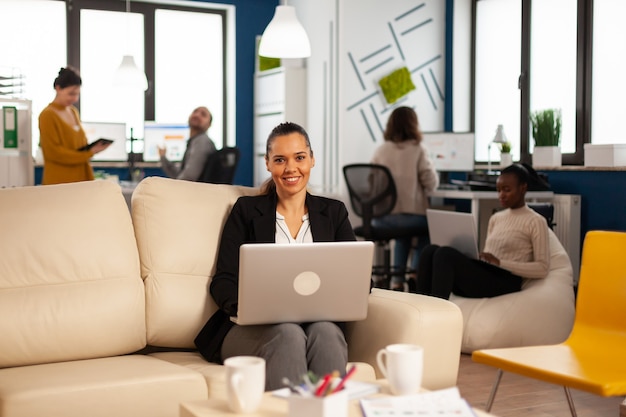 Mujer de negocios hispana sonriendo a la cámara sentado en el sofá escribiendo en la computadora portátil mientras diversos colegas trabajan en segundo plano.