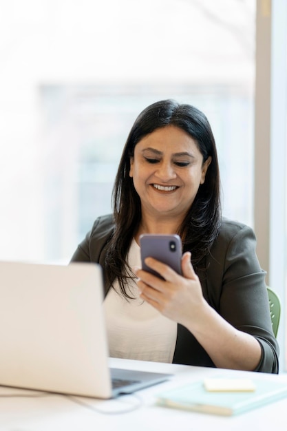 Mujer de negocios india usando una computadora portátil sosteniendo un teléfono móvil revisando el correo electrónico sentada en una oficina moderna