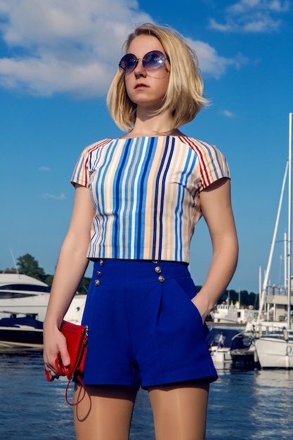 Mujer de negocios joven en baúles azules cortos de pie y esperando una reunión en el muelle con yates y barcos