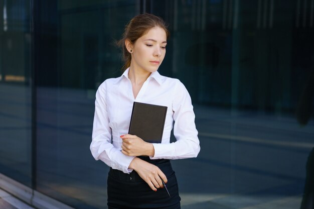 Mujer de negocios joven con una camisa blanca con un bloc de notas en la mano cerca de un edificio de oficinas. Concepto de negocio