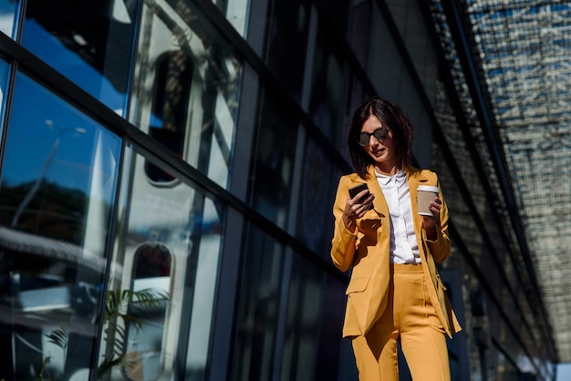 Mujer de negocios joven caucásica con smartphone y bebe café fuera cerca del centro de la oficina.