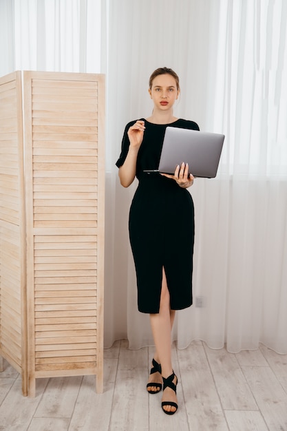 Mujer de negocios joven con la computadora en sus manos en vestido negro en la oficina blanca