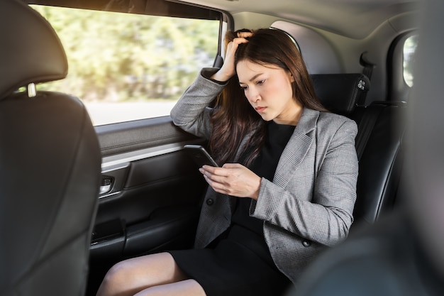 Mujer de negocios joven estresada que usa un teléfono inteligente mientras está sentado en el asiento trasero del coche