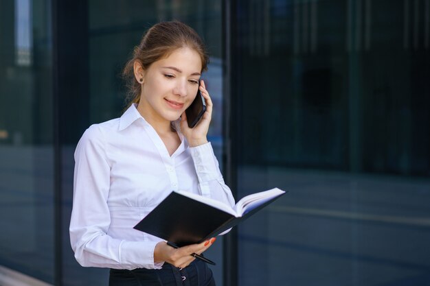 Mujer de negocios joven hablando por teléfono y escribiendo en un cuaderno. Con una camisa blanca de pie en el edificio de oficinas