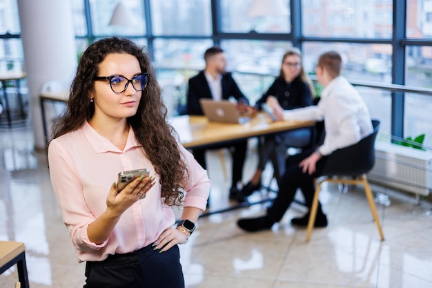 Mujer de negocios joven inteligente y feliz, con ropa y gafas elegantes, trabaja en la oficina, habla con un cliente en un teléfono inteligente, gestos, sonrisas. Los empleados están trabajando en segundo plano. enfoque selectivo