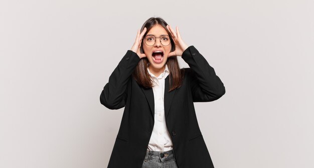 Mujer de negocios joven levantando las manos a la cabeza, con la boca abierta, sintiéndose extremadamente afortunada, sorprendida, emocionada y feliz