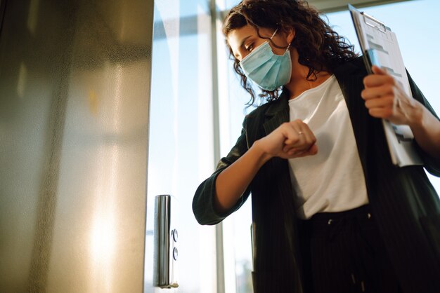 Mujer de negocios joven en máscara protectora utiliza un codo para presionar el botón del ascensor.