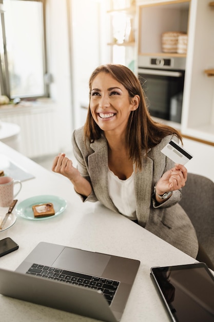 Mujer de negocios joven multitarea que usa una computadora portátil y compra en línea en su cocina por la mañana mientras se prepara para ir a trabajar.