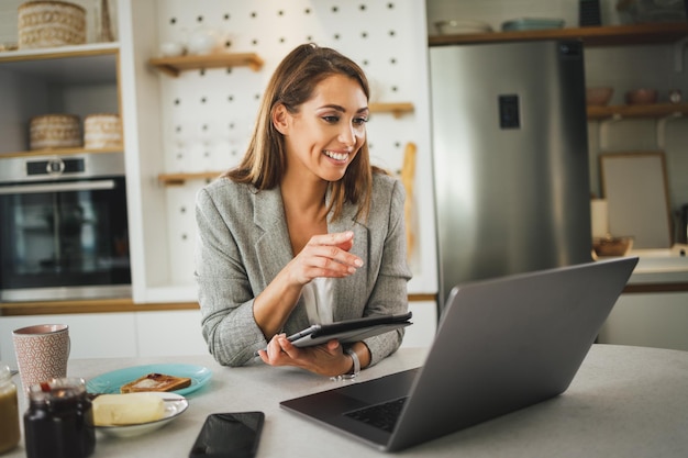 Mujer de negocios joven multitarea que usa una computadora portátil y una tableta digital y prepara un desayuno en su cocina.