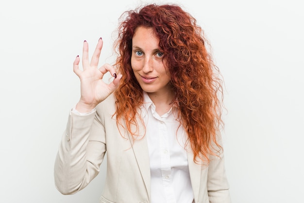 Mujer de negocios joven pelirroja natural aislada contra blanco alegre y confidente mostrando gesto bien.