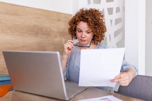 Mujer de negocios joven del pelo rizado con los vidrios que miran documentos en su mano mientras que se sienta en su oficina con la computadora portátil abierta. Mujer que trabaja en la oficina usando una computadora portátil, gerente femenina revisando documentos