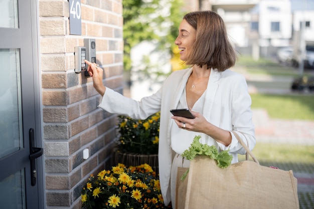 Mujer de negocios joven que va a casa con comestibles, ingresa una contraseña en el teclado para acceder. Concepto de tecnologías modernas en la vida cotidiana