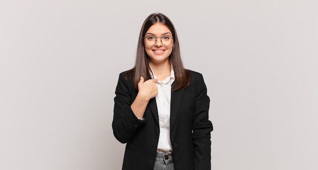 Mujer de negocios joven que parece feliz, orgullosa y sorprendida, apuntando alegremente a sí misma, sintiéndose segura y elevada